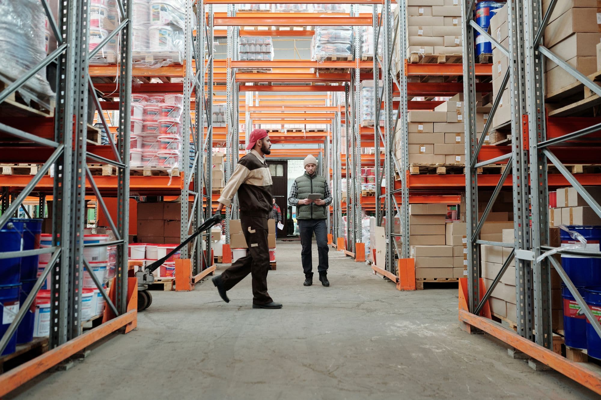 Warehouse aisle with a worker moving a pallet and a supervisor holding a tablet
